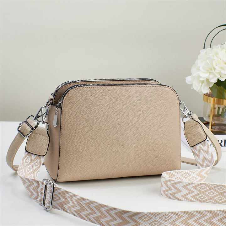 Beige leather crossbody bag with double zipper and adjustable patterned strap on white surface