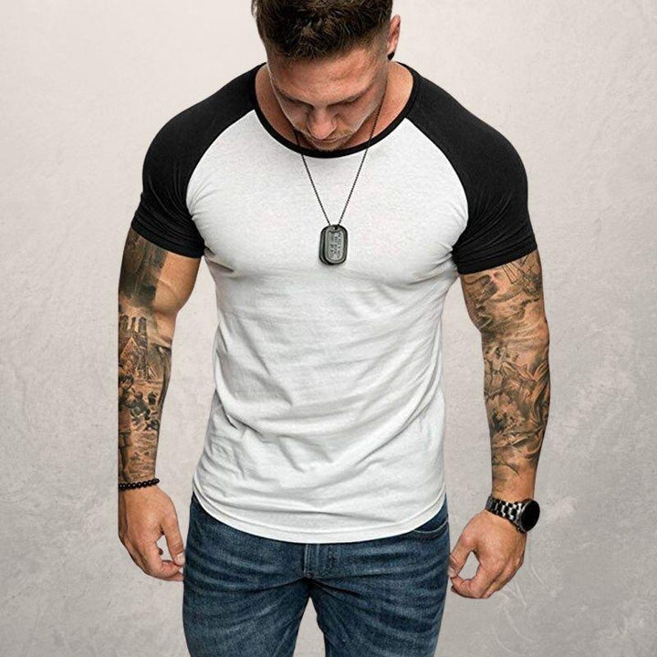 muscular man wearing black and white raglan t-shirt with tattooed arms, necklace, watch, and jeans against gray background