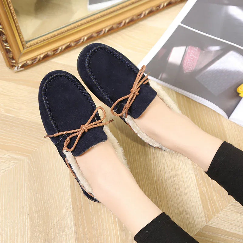 Navy blue suede slip-on loafers with brown bow laces and beige fur lining worn on feet on wooden floor