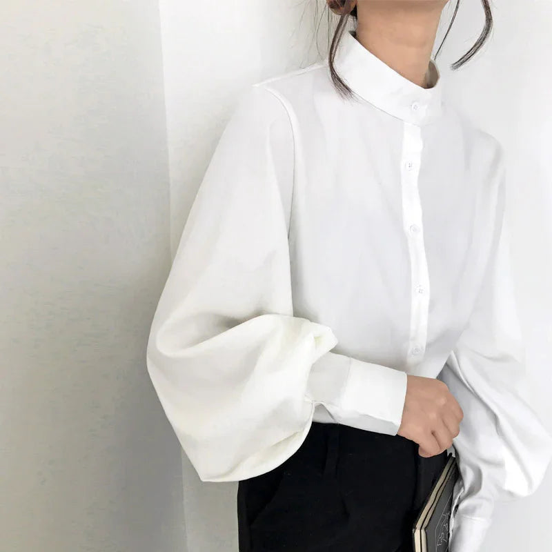 Woman wearing a white puff sleeve blouse with a mandarin collar and black pants against a plain wall