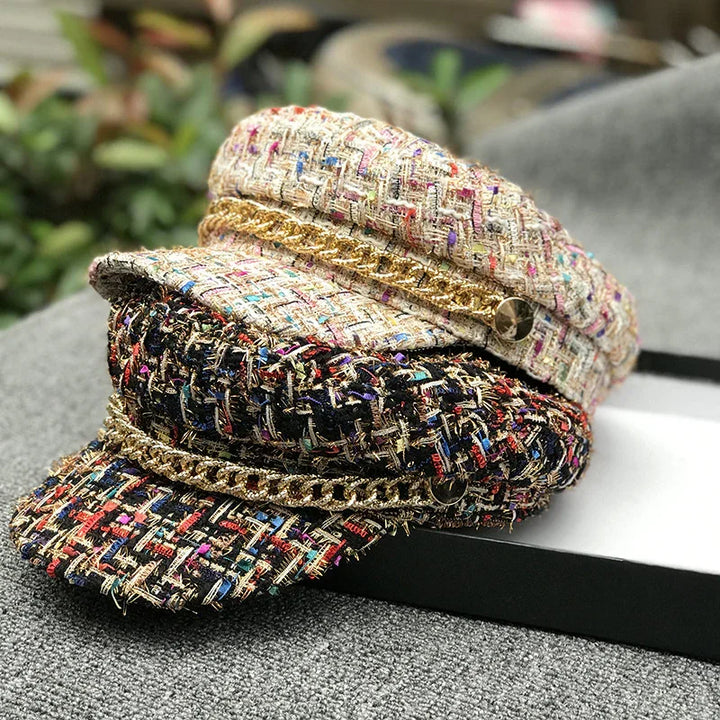 Stack of two textured tweed baker boy hats with gold chain detail, one beige and one black multicolor, on gray fabric surface