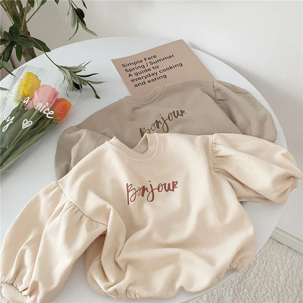 Beige and light brown puff-sleeve sweatshirts with 'Bonjour' text on white round table with flowers and book