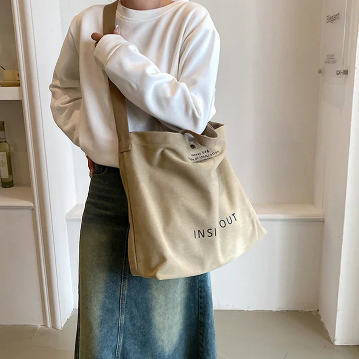 Person wearing white sweatshirt and denim skirt carrying beige canvas tote bag with 'INSI OUT' text