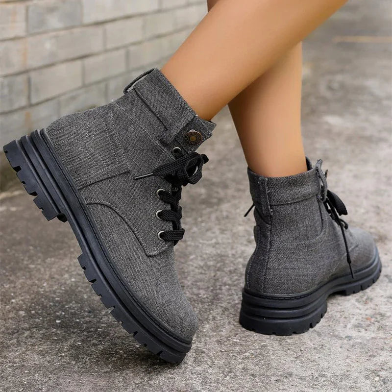 Gray fabric lace-up ankle boots with black thick soles worn on concrete pavement near a brick wall
