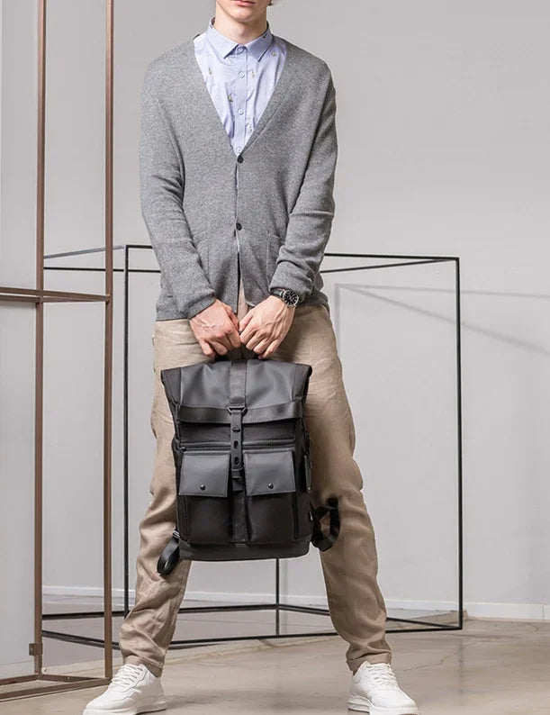 Man wearing gray cardigan and beige pants holding black leather backpack in minimal modern room