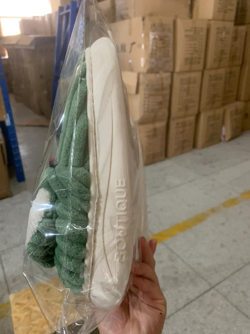 Hand holding packaged green and beige BOUTIQUE brand slippers in a warehouse with stacked cardboard boxes