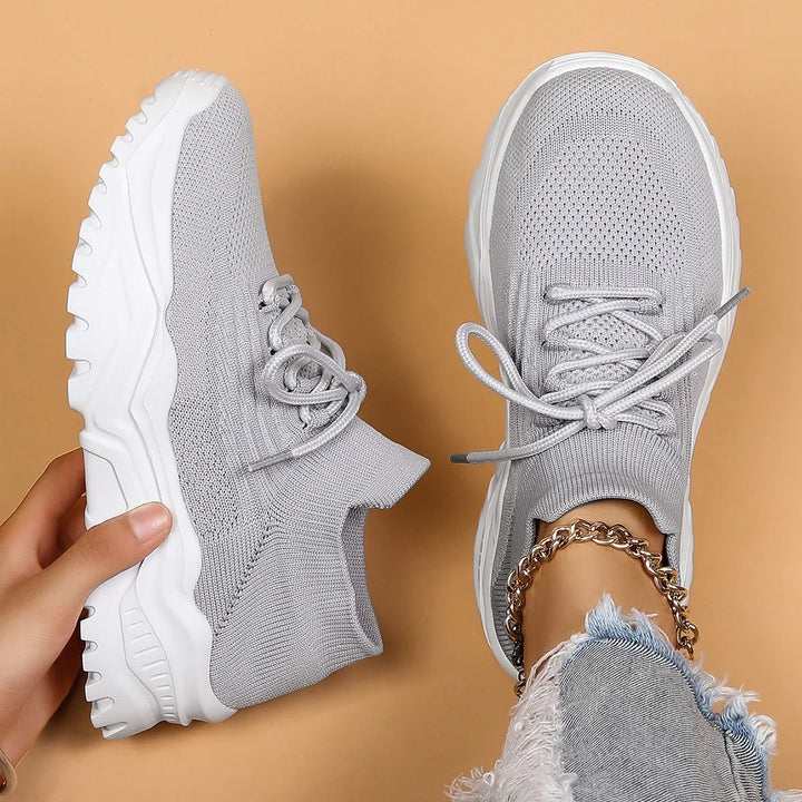Light gray knit sneakers with white chunky soles on beige background, one worn with gold ankle chain