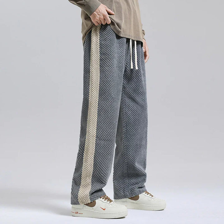 Man wearing textured gray sweatpants with beige side stripe and white sneakers on gray background