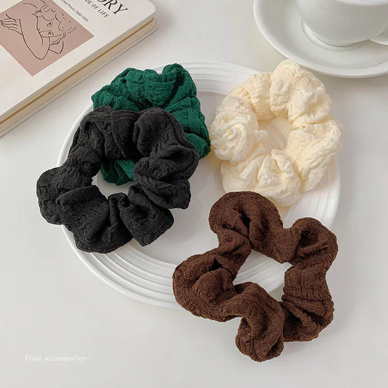 Set of four textured fabric hair scrunchies in green, black, cream, and brown on a white plate