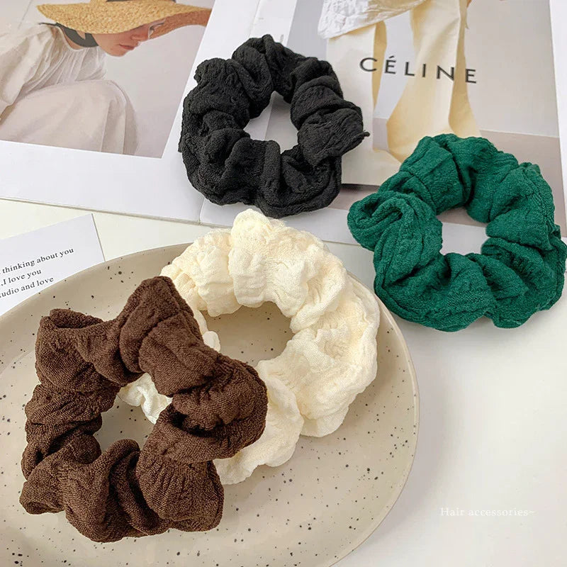 Set of four textured fabric scrunchies in black, green, brown, and cream on speckled plate with fashion magazines background
