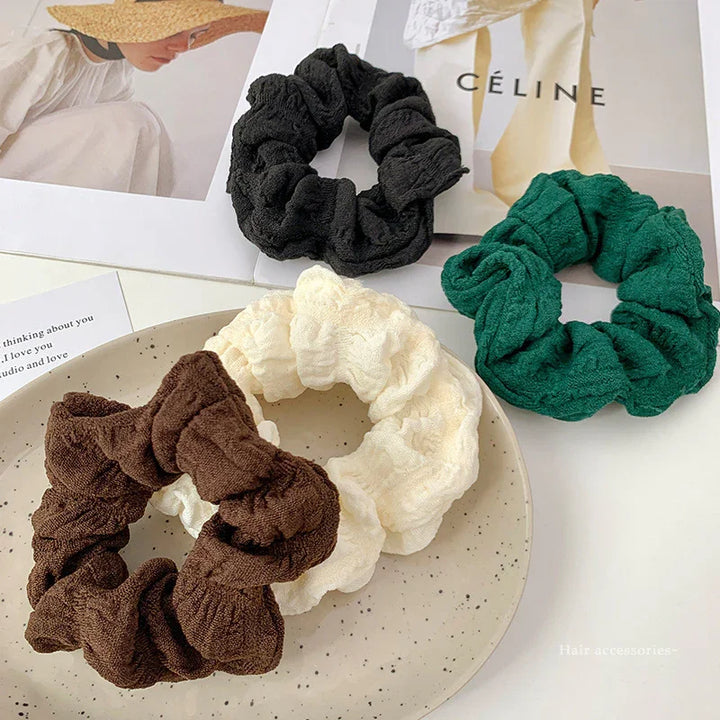 Set of four textured fabric scrunchies in black, green, brown, and cream on speckled plate with fashion magazines background