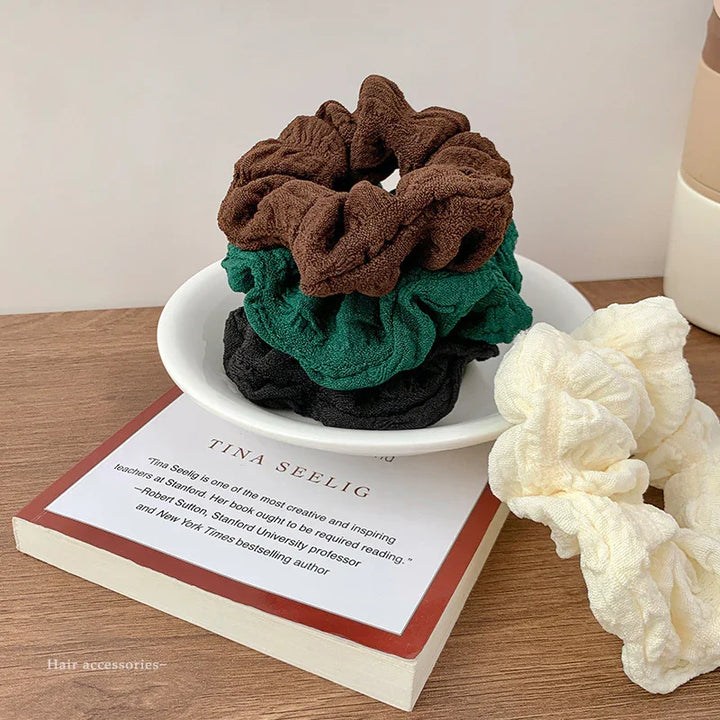 Stack of brown, green, and black scrunchies in a white dish with a cream scrunchie on wooden table next to book