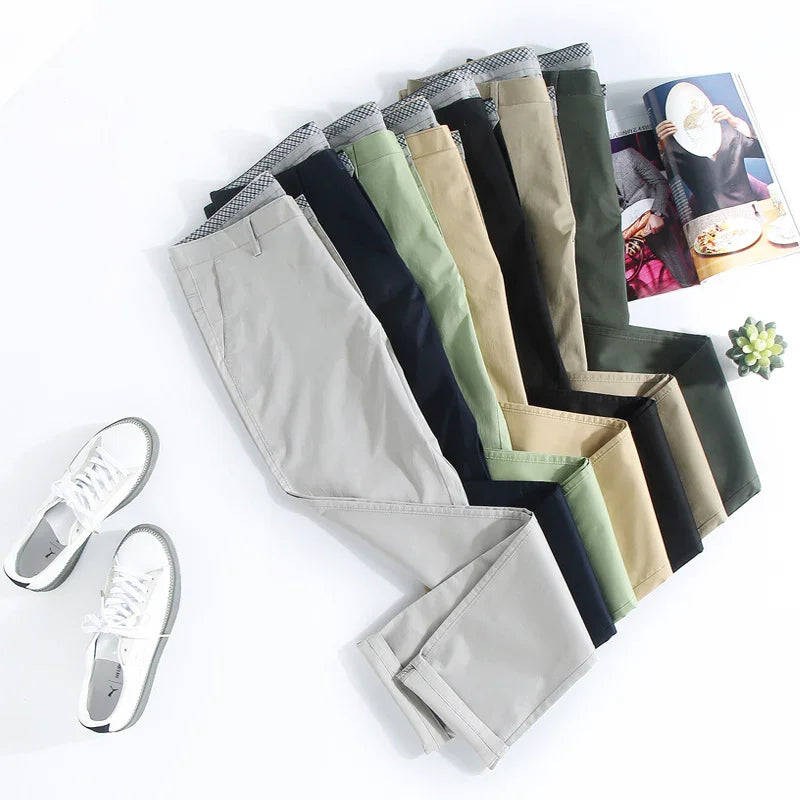 Seven pairs of casual men's pants in assorted colors neatly folded with white sneakers and an open magazine on white surface