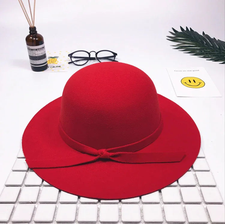 Red wool felt wide-brim hat with a bow detail on a white tiled surface with glasses, diffuser, and decorative cards