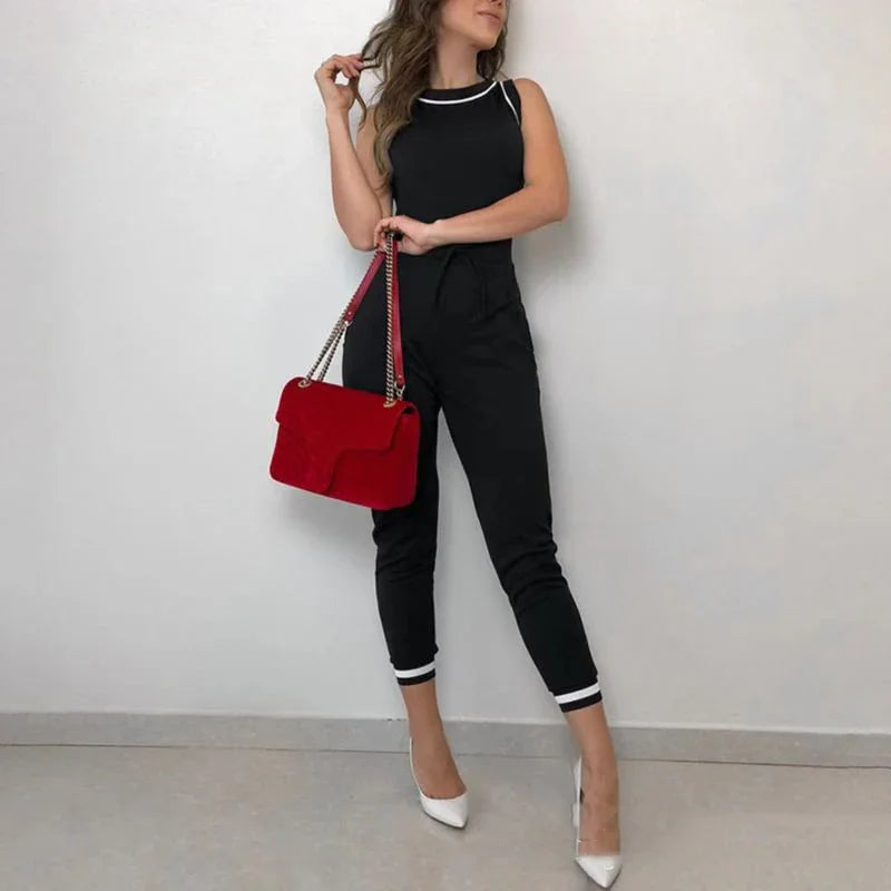 Woman in black sleeveless jumpsuit holding red handbag, wearing white pointed heels, standing against plain wall