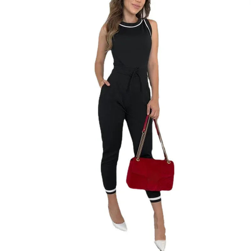 Woman wearing black sleeveless jumpsuit with white trim, holding red handbag and white heels
