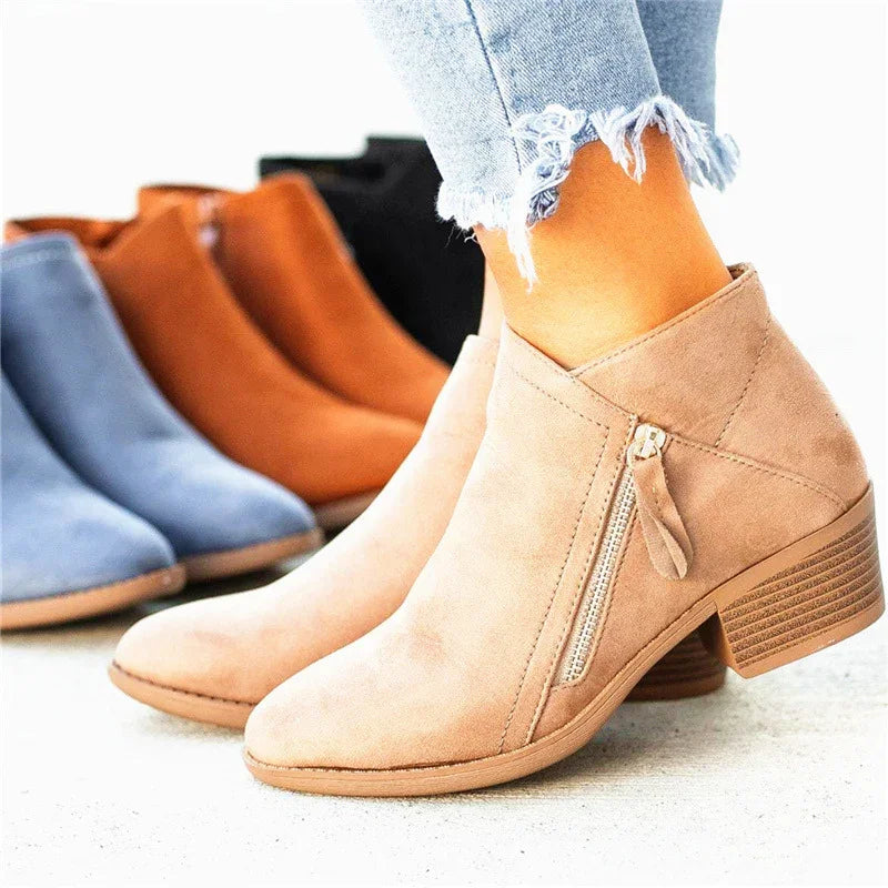 Women's suede ankle boots in beige with zipper detail, worn with frayed light blue jeans, other boots in blue, brown, and black in background