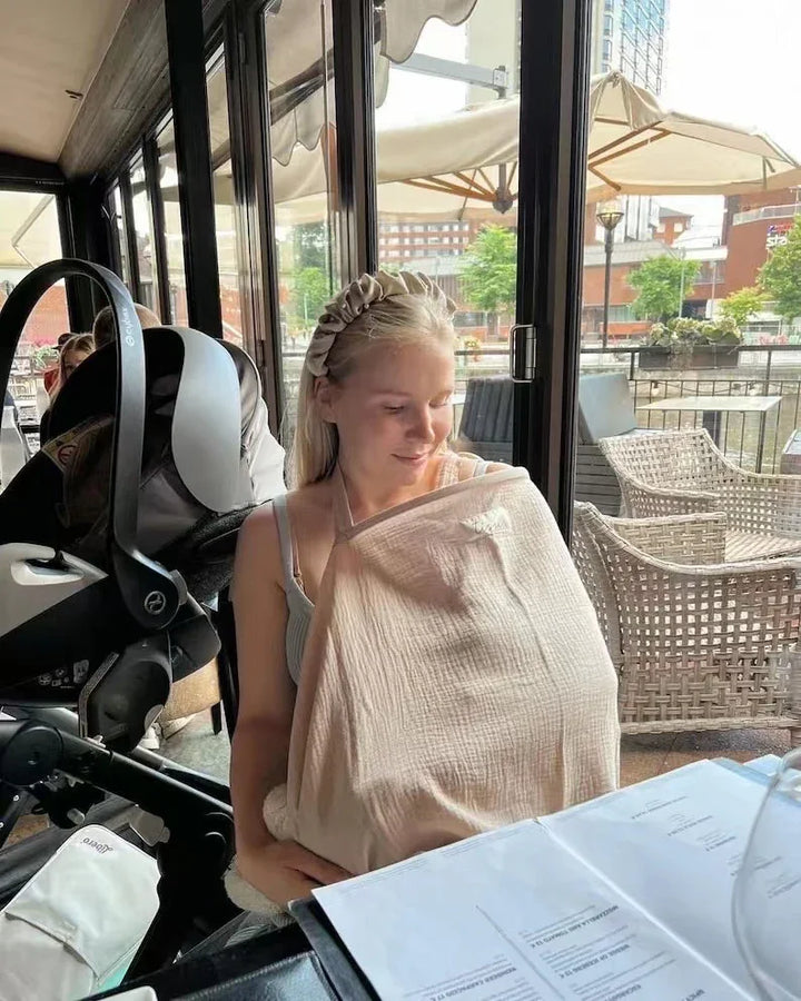 Young mother nursing baby under a beige cover at outdoor café with stroller beside her
