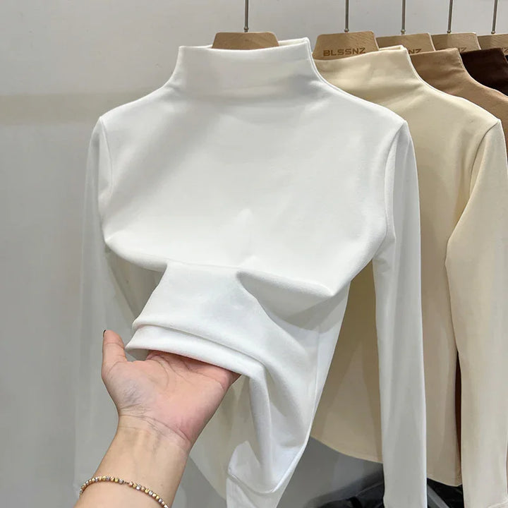 White long sleeve turtleneck shirt displayed on wooden hanger with hand holding fabric, beige and brown shirts in background