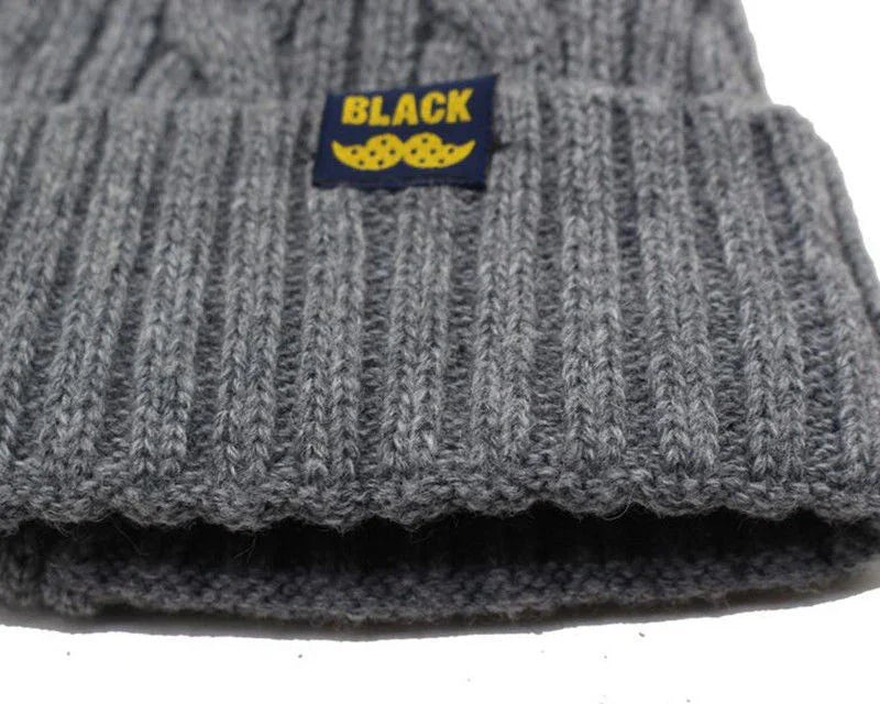 Close-up of gray ribbed knit beanie with folded cuff and black label featuring yellow text 'BLACK' and yellow mustache design
