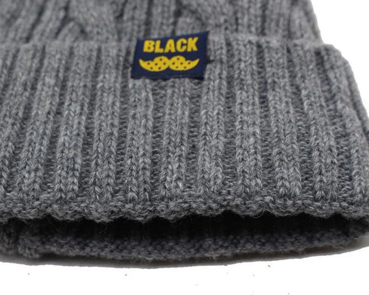 Close-up of gray ribbed knit beanie with folded cuff and black label featuring yellow text 'BLACK' and yellow mustache design