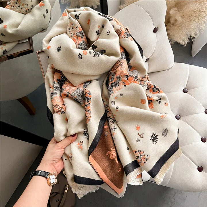 Hand holding a cozy beige scarf with orange and black floral patterns on a tufted cream chair