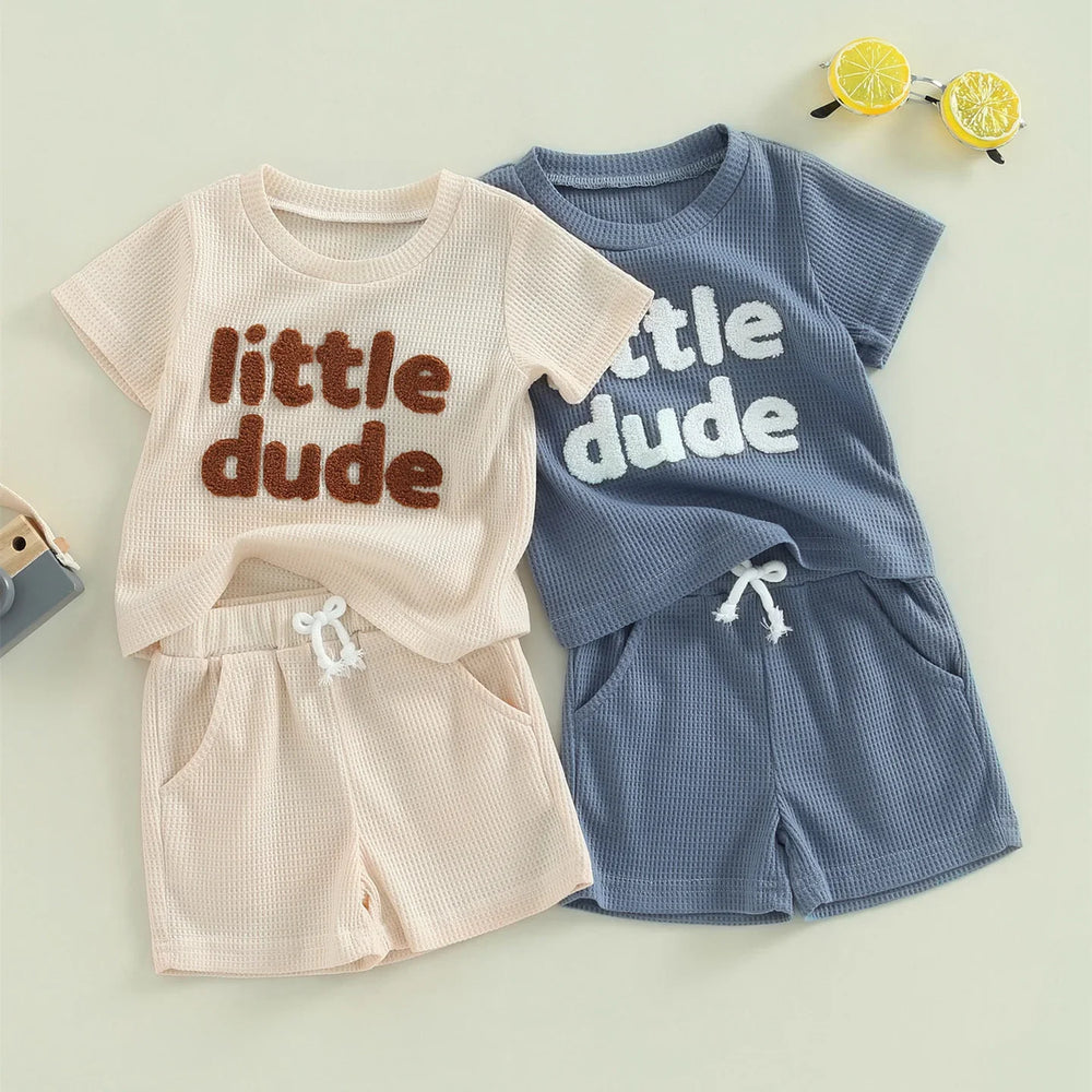 Two textured baby outfits with shorts and t-shirts in beige and blue, printed 'little dude'