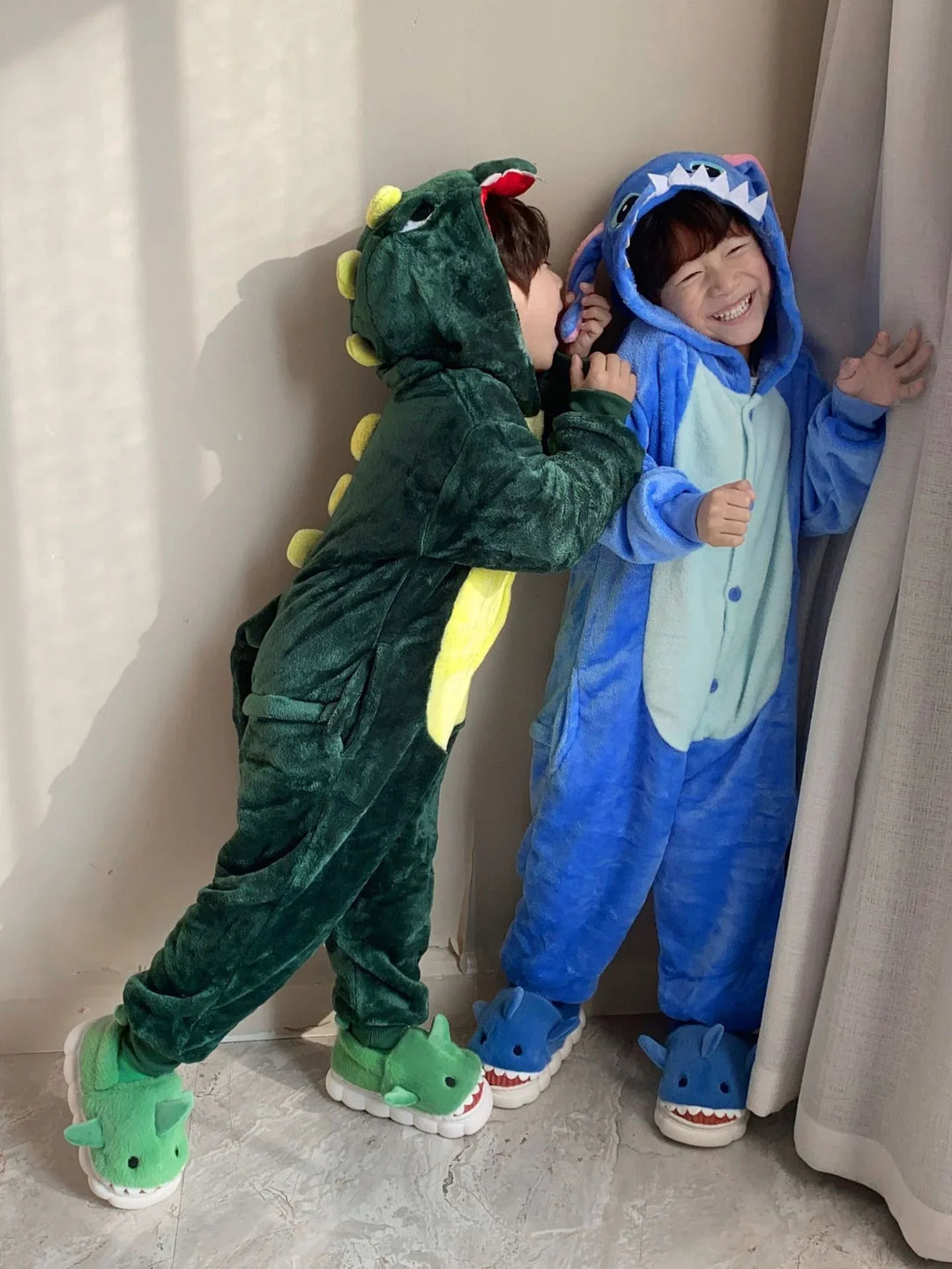 Two children playing indoors wearing green dinosaur and blue shark plush onesie pajamas with matching slippers