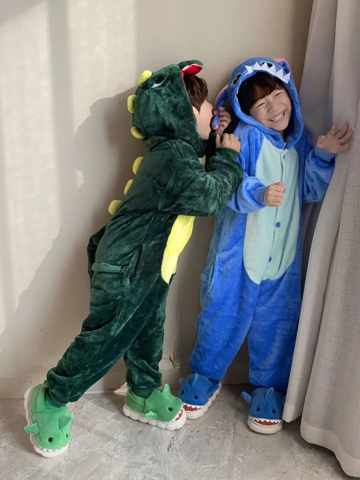 Two children playing indoors wearing green dinosaur and blue shark plush onesie pajamas with matching slippers