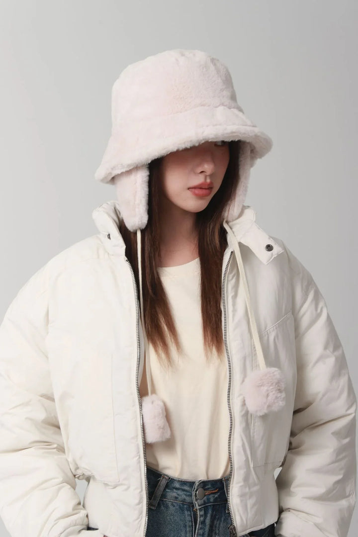 Woman wearing white fuzzy bucket hat with earflaps and pom-poms, white puffer jacket, and casual outfit