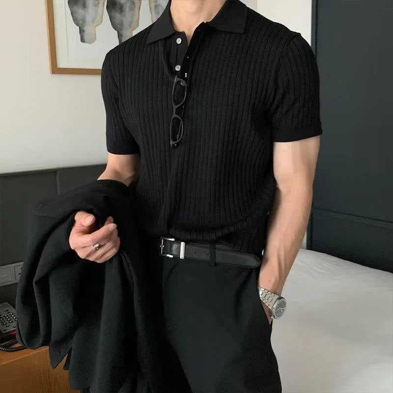Man wearing black ribbed polo shirt with black pants and belt, holding black jacket indoors