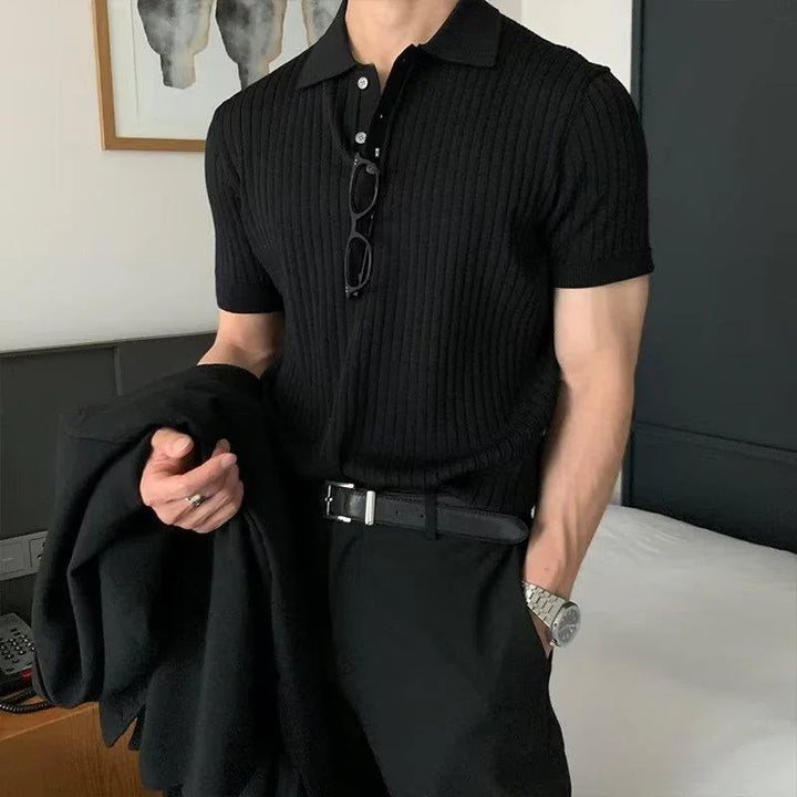 Man wearing black ribbed polo shirt with black pants and belt, holding black jacket indoors