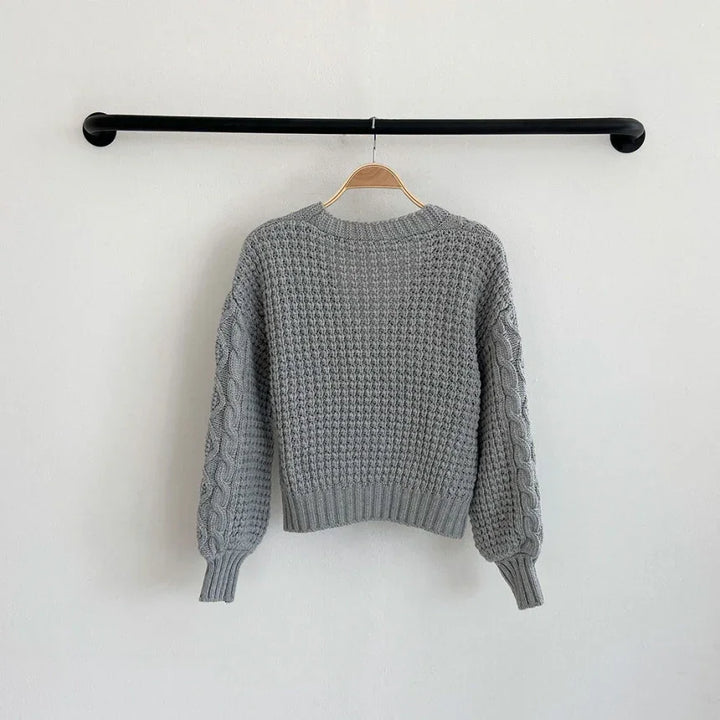 Gray knitted sweater with textured front and cable knit sleeves hanging on wooden hanger against white wall