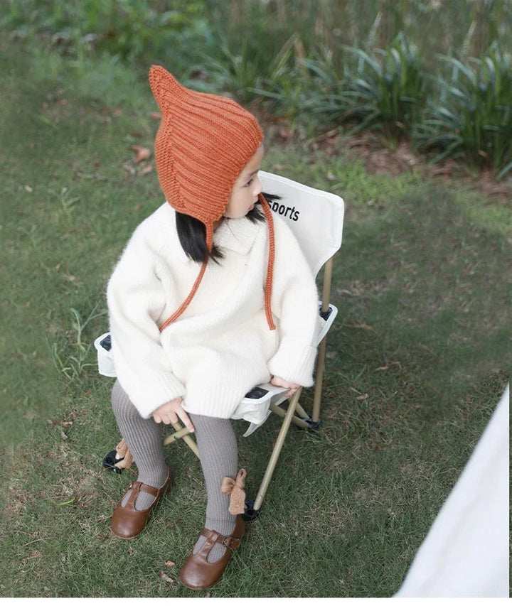 Toddler girl wearing an orange knit hat, white sweater dress, and brown shoes sitting on a small outdoor chair on grass