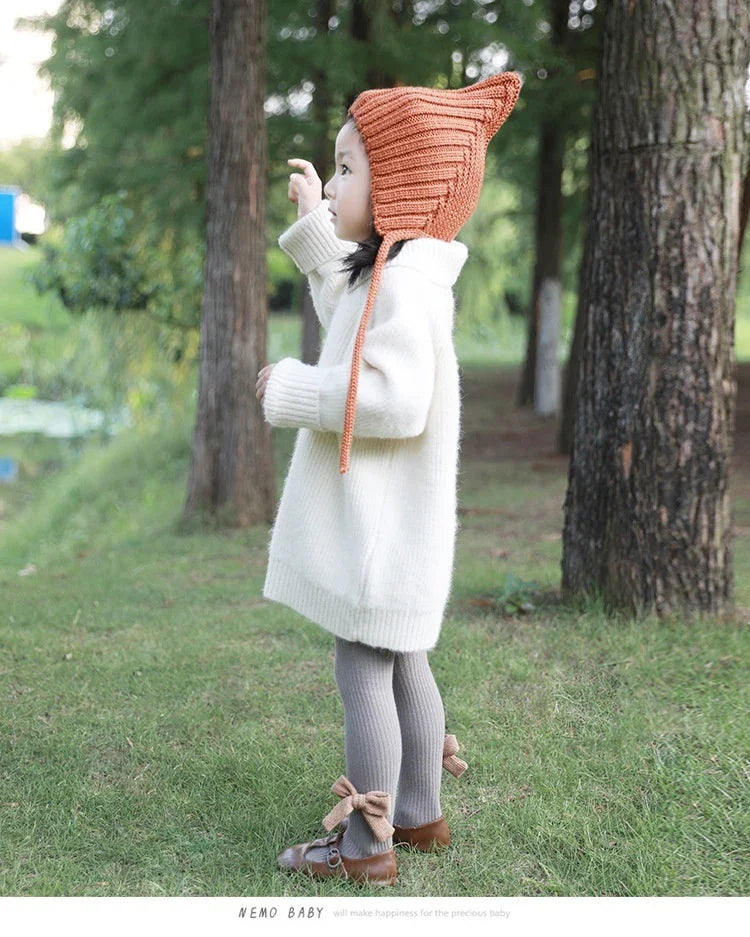 Toddler girl in white sweater dress and orange knit hat standing in forest park with grass and trees