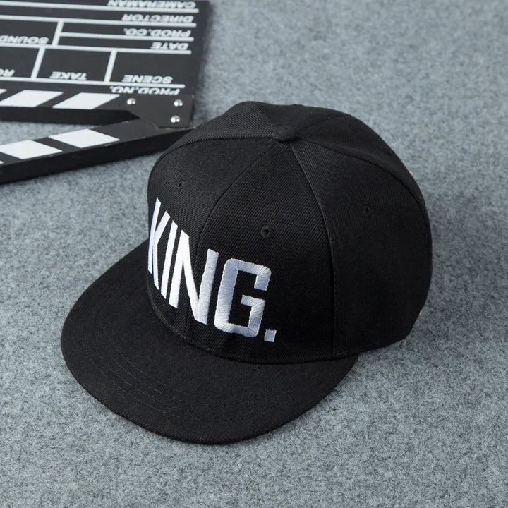 Black snapback cap with white embroidered text 'KING.' on front panel, placed on gray surface with film clapperboard nearby