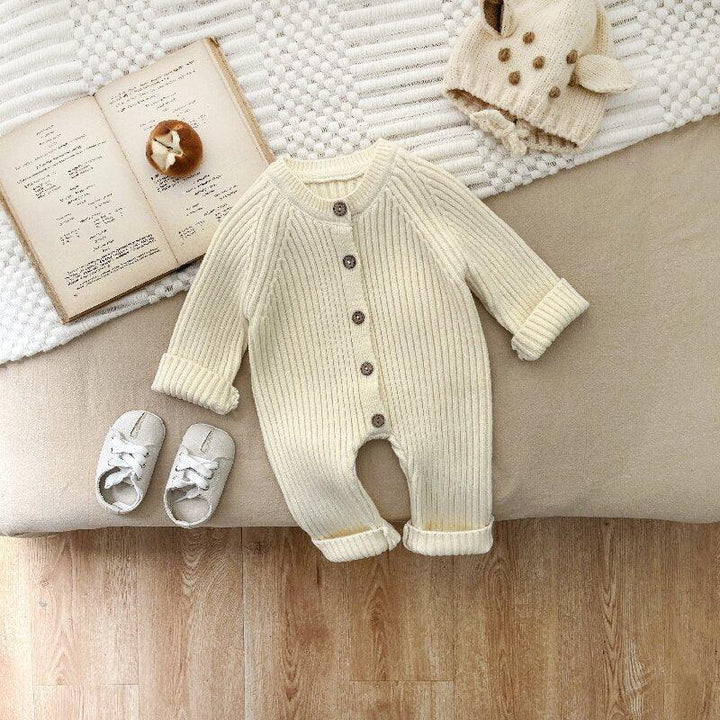 Cream ribbed baby romper with button closure, white soft baby shoes, knit animal hat, and open book on beige and white textured background