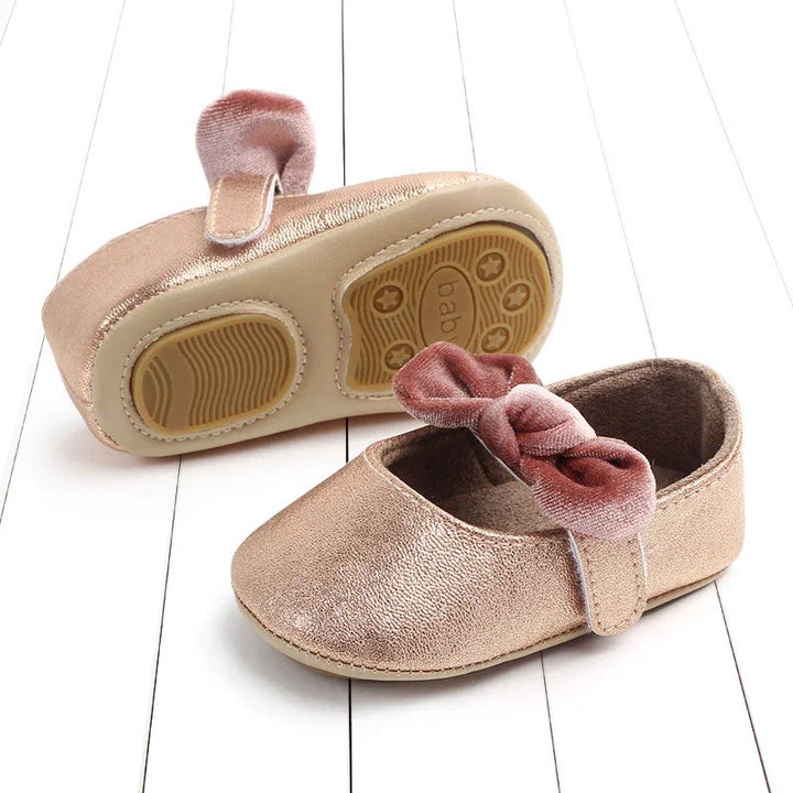 Pair of metallic rose gold baby shoes with velcro strap and pink velvet bow on white wooden floor