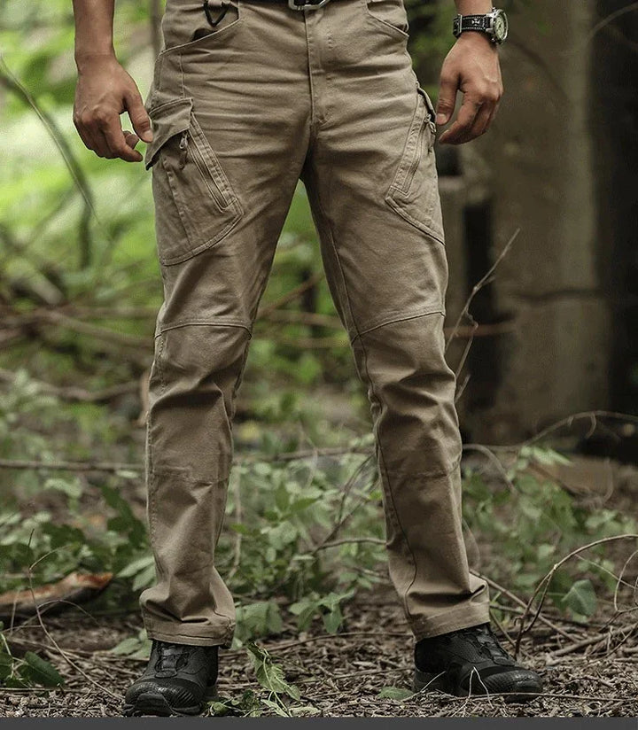 Man wearing beige cargo pants and black shoes standing outdoors on foliage-covered ground