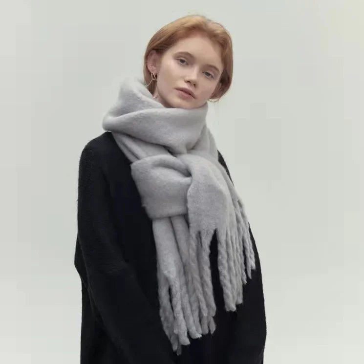 Young woman with red hair wearing a large light gray fringed scarf and black sweater on light background