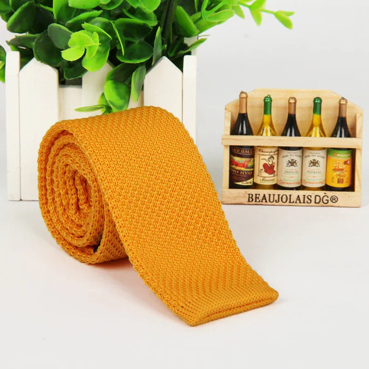 Rolled mustard yellow knitted tie displayed with green plant in white picket fence and miniature wine bottles in wooden crate
