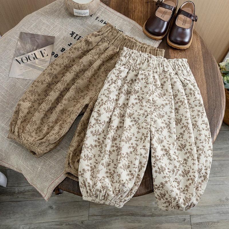 Two pairs of floral print children's pants in beige and white on wooden table with brown shoes and Vogue magazine