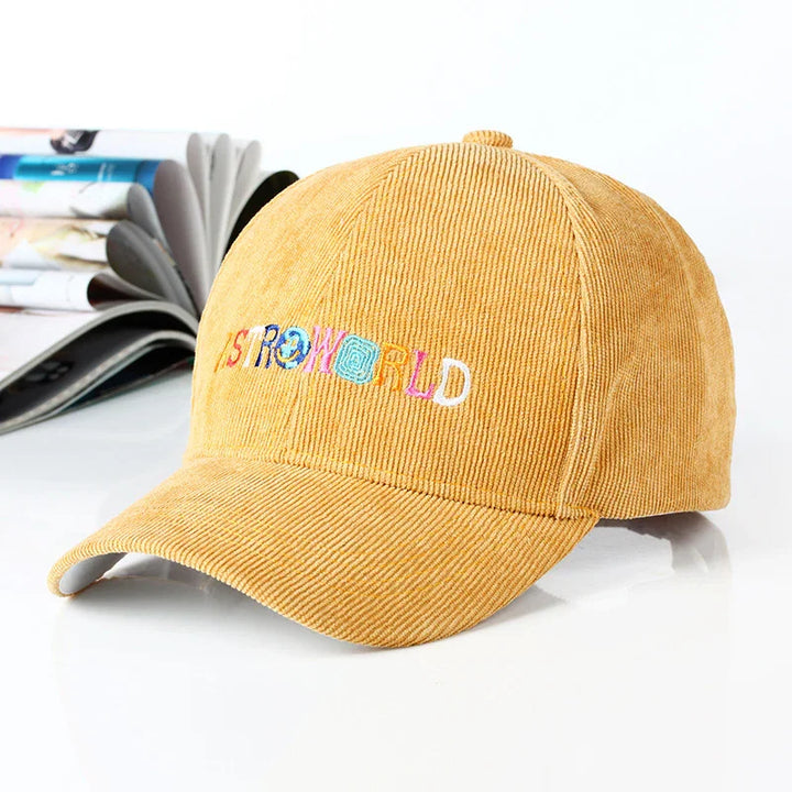 Yellow corduroy baseball cap with colorful embroidered Astroworld logo on front, open magazines in background