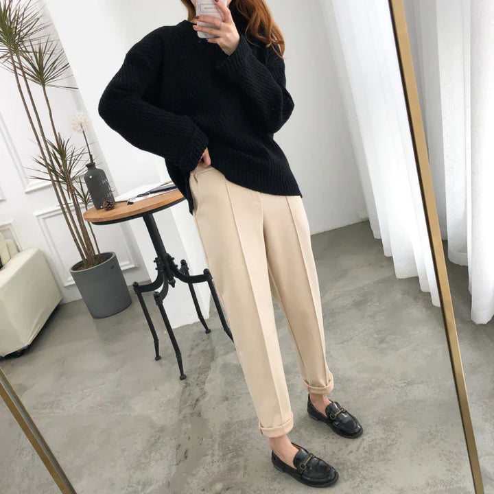 Woman wearing black knit sweater, beige pleated pants, and black loafers taking mirror selfie in modern room