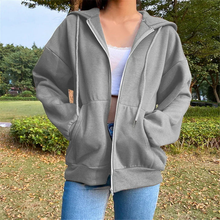 Woman wearing a grey zip-up hoodie with pockets and drawstrings, paired with blue jeans, outdoors in a park