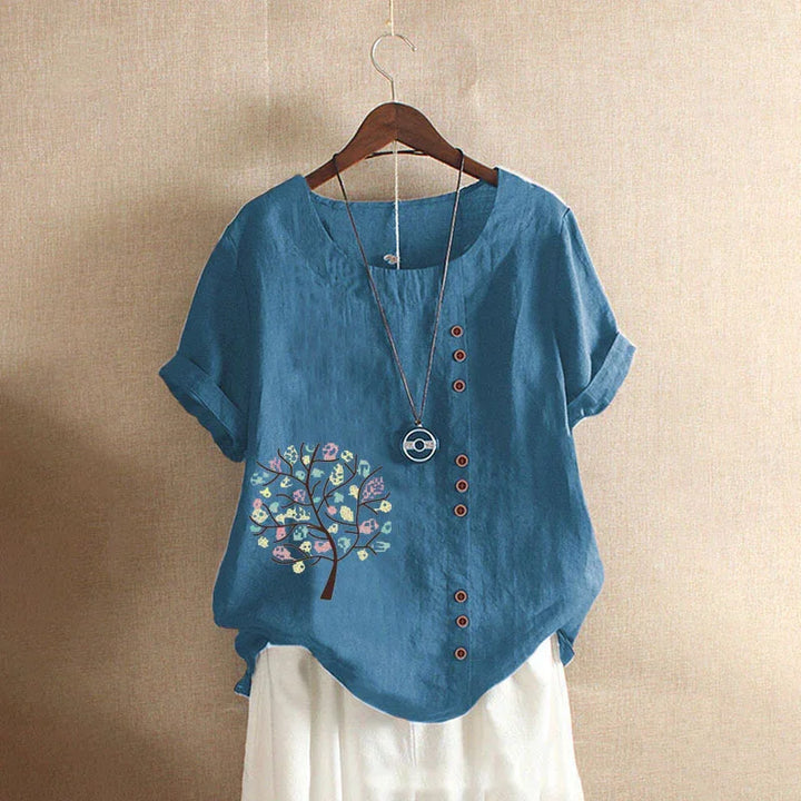 Blue short-sleeve linen blouse with multicolor tree print and decorative buttons, displayed on wooden hanger