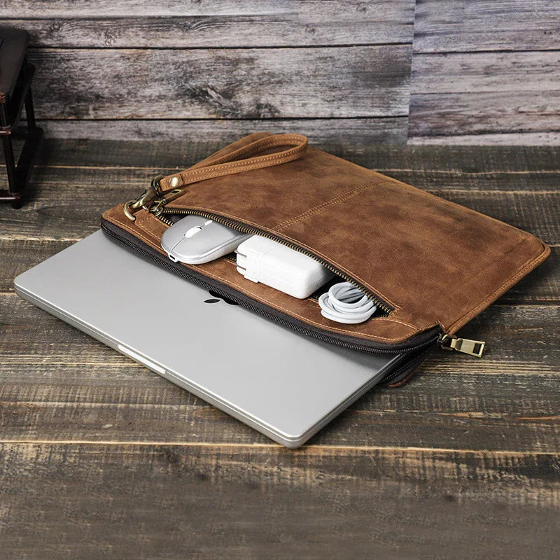 Brown leather laptop sleeve with zipper holding silver laptop, mouse, charger, and cable on wooden surface