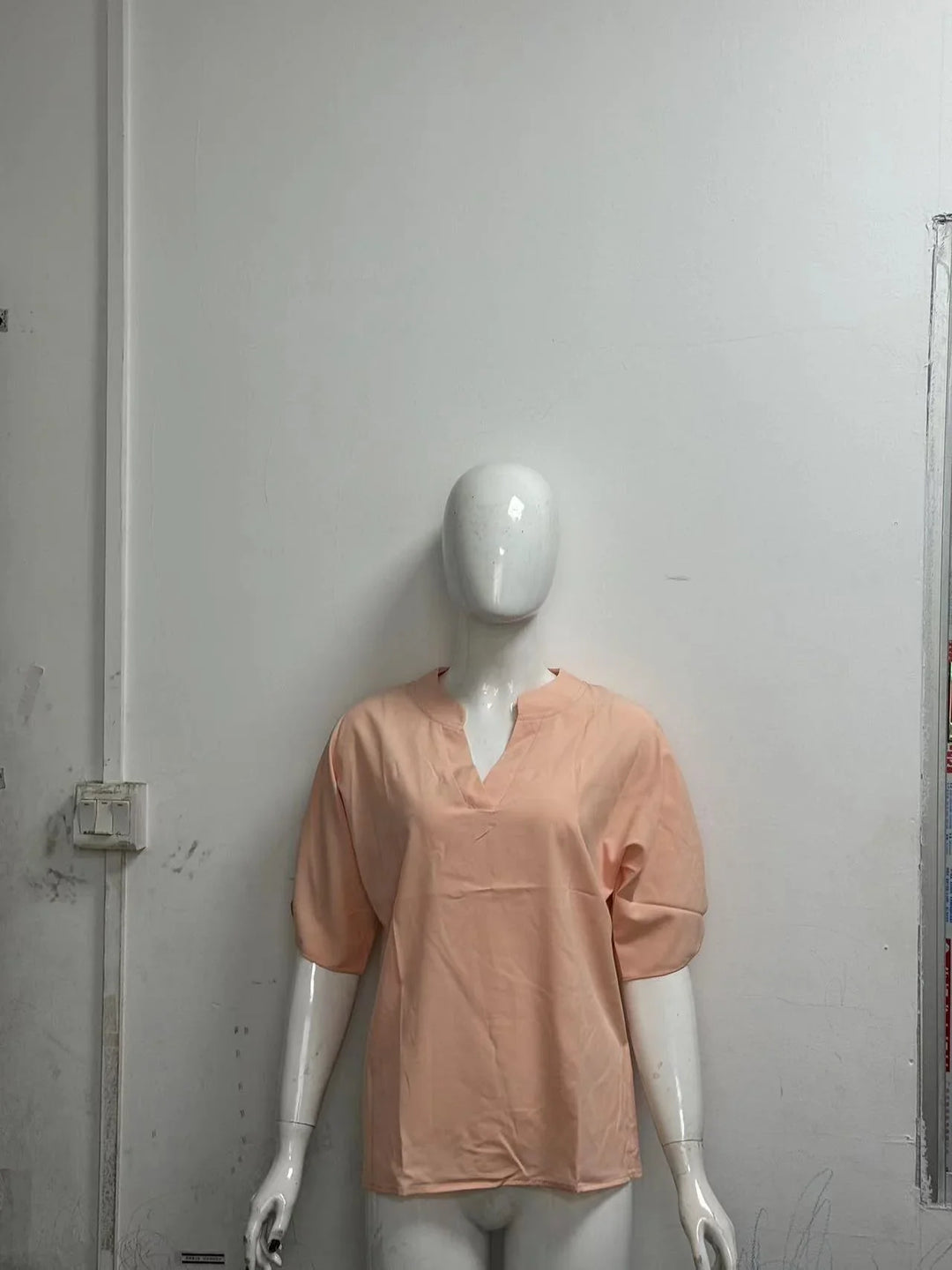 White mannequin wearing a peach short-sleeve blouse with a V-notch neckline against plain gray wall