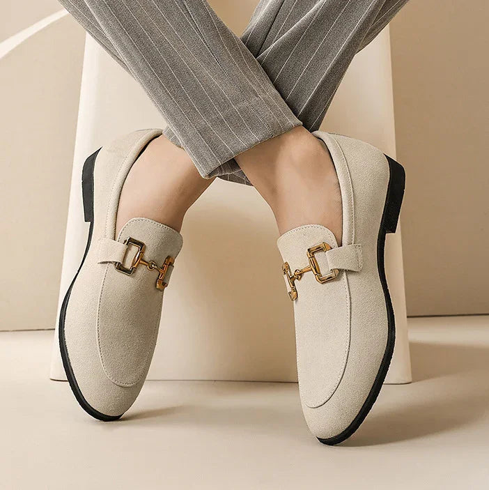 Beige suede loafers with gold buckle detail worn with gray pinstripe trousers on beige background