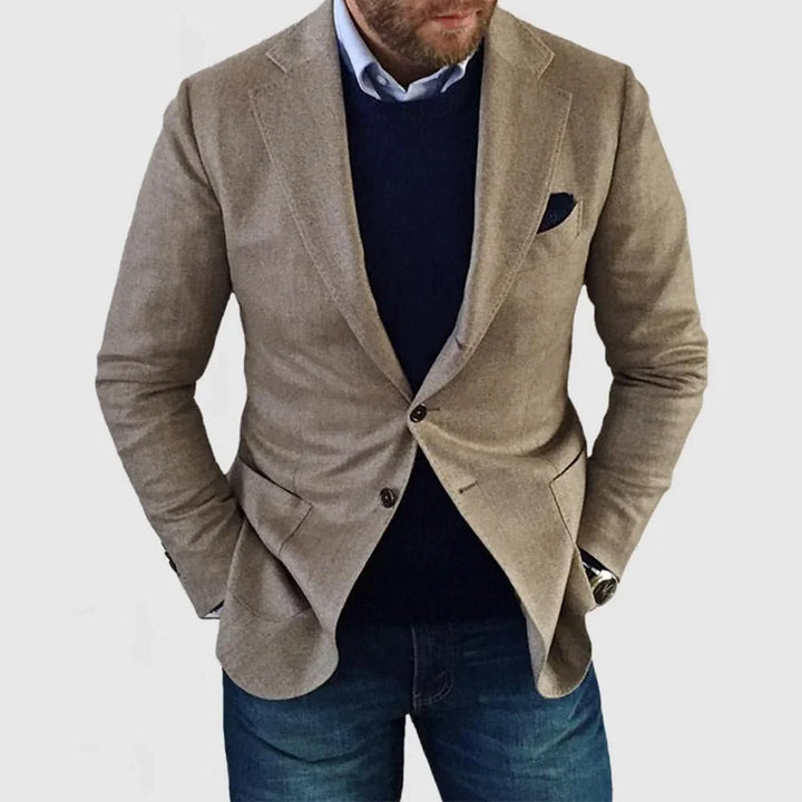 Man wearing beige blazer over navy sweater and blue jeans against white background